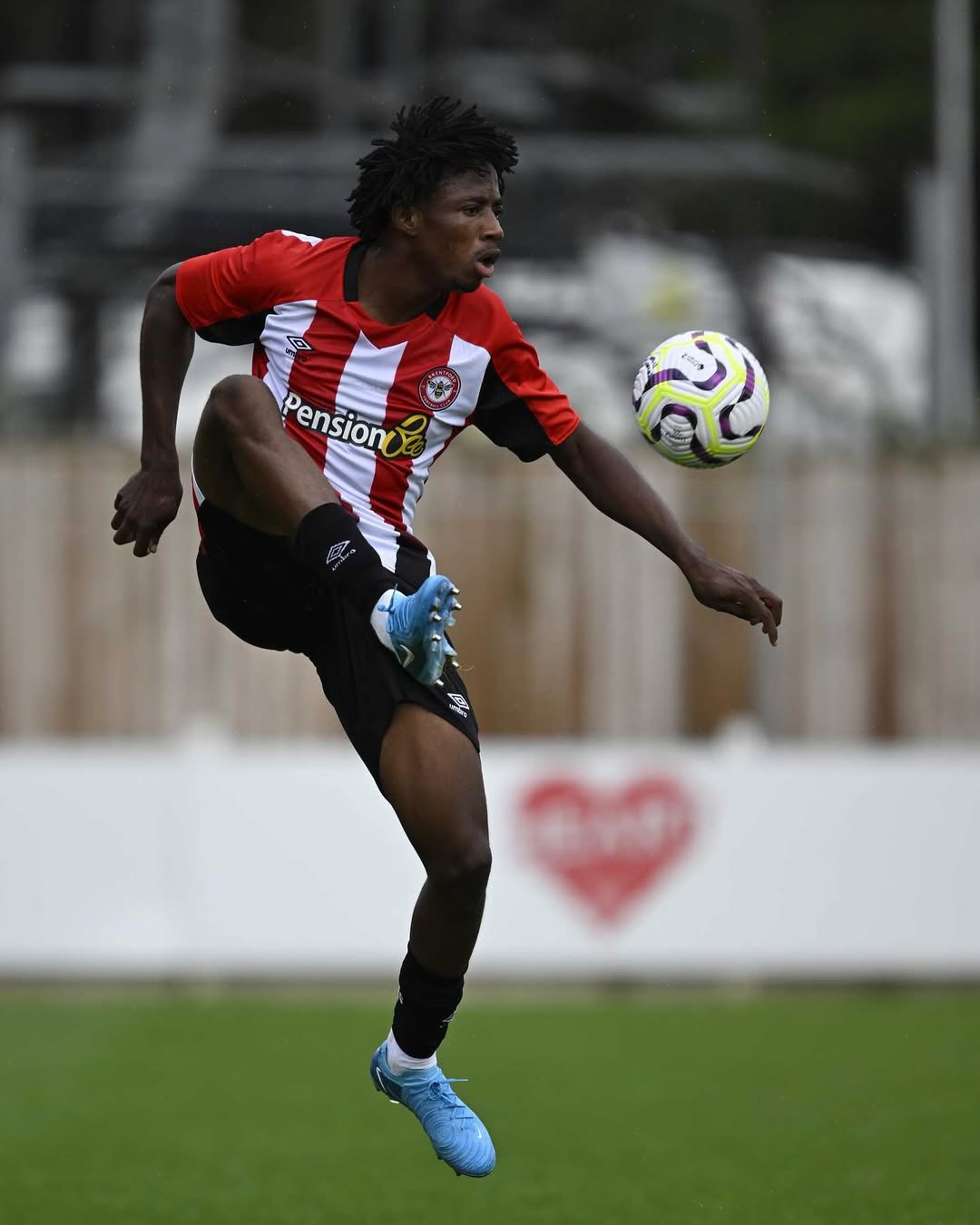 Benjamin Fredrick controls the ball in mid-air, wearing Brentford B's red and white striped kit during a match